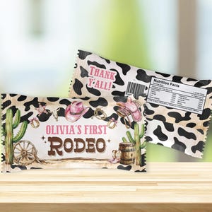 May include: Two candy wrappers with a cowhide pattern and rodeo-themed graphics. One wrapper reads "Olivia's First Rodeo" with a cactus, a pink cowboy hat, and a barrel. The other wrapper says "Thank Y'all!" and includes a nutrition facts label.
