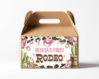 May include: A brown cardboard favor box with a handle. The box is decorated with a cowhide pattern and a pink cowboy hat. The text "Olivia's First Rodeo" is printed on the front of the box.