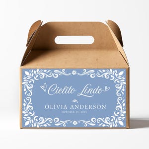 Könnte beinhalten: Eine braune Geschenkbox aus Pappe mit Griff. Die Vorderseite der Box zeigt ein hellblaues rechteckiges Etikett mit weißem Blumen- und Scrollwork-Rand. Auf dem Etikett steht "Cielito Lindo", "Olivia Anderson" und "25. Oktober 2023".