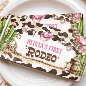 May include: Two rectangular party favor boxes with a Western theme. The boxes feature a cowhide pattern, pink cowboy hats, saddles, and the text "Olivia's First Rodeo." Decorative elements include cacti, barrels, and a sheriff's badge.