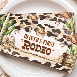 Può includere: Una scatola di bomboniere per feste a tema cowboy, di colore marrone e bianco, con un motivo di pelle di mucca marrone e bianco. La scatola ha il testo "Oliver's First Rodeo" stampato in lettere marroni.