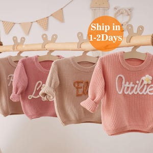 May include: Four pastel-coloured knit sweaters hang on a wooden rod. Each sweater has a different name embroidered on the front in white. The sweaters are in shades of pink and beige. A wooden teepee and banner are in the background.