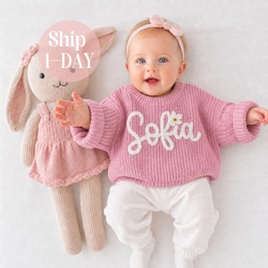 Peut inclure: Un bébé portant un pull en tricot rose avec le prénom Sofia brodé en écriture blanche et une petite fleur blanche, accompagné d'un pantalon blanc et d'un bandeau rose. À côté se trouve une poupée lapin en tricot rose portant une robe.
