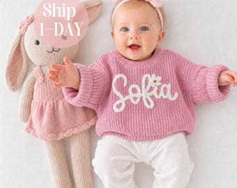 Könnte beinhalten: Ein Baby trägt einen rosa Strickpullover mit dem in weißer Schreibschrift gestickten Namen Sofia und einer kleinen weißen Blume, dazu eine weiße Hose und ein rosa Haarband. Daneben liegt eine passende rosa Strick-Hasenpuppe mit Kleid.