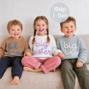 May include: Three children wearing long-sleeve ribbed knit jumpers. The left jumper is tan with blue text reading 'lil bro', the centre is white with purple text reading 'big sister', and the right is light blue with white text reading 'big bro'.
