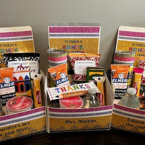 May include: Three gift baskets filled with school supplies and treats. Each basket has a gold glitter base with pink and silver stripes. The baskets contain composition books, snacks, and water bottles. Text on the baskets reads "It Takes a Big Heart to Teach" and "Educators are the real MVPs!"