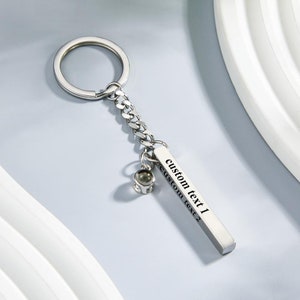 May include: Silver keyring with a silver chain and a rectangular charm with the text "custom text 1" and "custom text 2" engraved on it. The charm has a small silver bead attached to it.