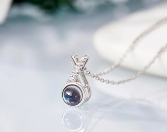 May include: A silver pendant necklace with an X-shaped design. The pendant has a round, dark-coloured centre and is accented with small, clear gemstones. The necklace chain is delicate and silver, reflecting light.