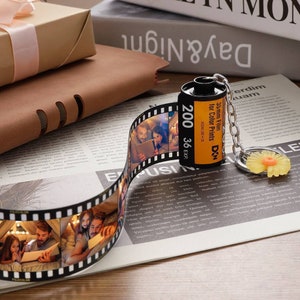 May include: A black and yellow film canister keychain with a silver chain. The canister has the text "35mm Film for Color Prints DIN" and "200 36 EXP" printed on it. The keychain is lying on a newspaper with a strip of film with photos printed on it.