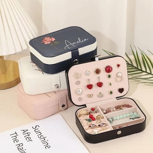 May include: Three jewellery boxes in different colours and designs. The top box is blue with a white rose and the name "Amelia" written on it. The middle box is white with a floral design. The bottom box is pink. The bottom box is open and shows a variety of jewellery, including earrings, rings, and bracelets.