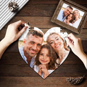 Peut inclure: Un puzzle en forme de cœur avec une photo d'une famille de trois personnes. Le puzzle est en carton blanc et a un fond blanc. La photo montre un homme, une femme et une jeune fille souriant à la caméra.