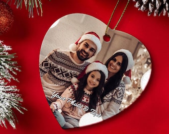 May include: Heart-shaped Christmas ornament featuring a family photo. The ornament has a gold string for hanging and the text "Your Photo & Text". The family is wearing Santa hats and sweaters. The background is red.