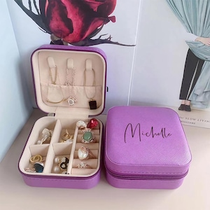 May include: Purple two-piece jewellery box with a zipper closure. The top box has a compartment for necklaces and the bottom box has compartments for rings and earrings. The bottom box has the name "Michelle" engraved on the top.