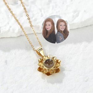 May include: A gold chain necklace with a gold flower pendant. The flower has a small round opening in the center that reveals a photo of two people.