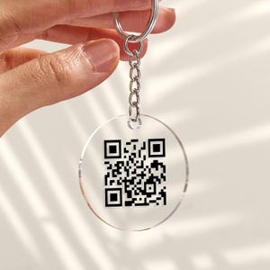 May include: A clear, circular keyring featuring a black QR code. The keyring is attached to a silver metal ring and chain. The QR code is a pattern of black and white squares.