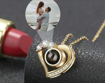 May include: A gold heart-shaped necklace with a small black circle in the centre. The circle projects an image when viewed through a small magnifying glass. The necklace is adorned with small clear rhinestones.
