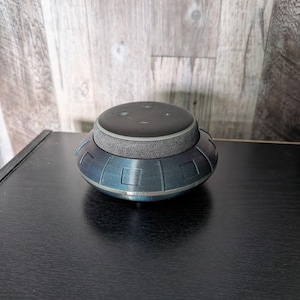 May include: A smart speaker sits atop a blue and silver decorative base. The base has a circular design with rectangular cutouts. The speaker is gray and black, and the base is on a black surface.