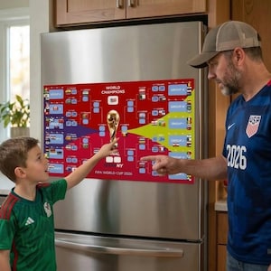 May include: A red poster on a refrigerator displays a gold trophy and the text "WORLD CHAMPIONS" and "FIFA WORLD CUP 2026 NY". A man and a boy are pointing at the poster. The boy wears a green jersey.