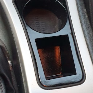 May include: A car's center console featuring a cup holder and a rectangular storage compartment. The cup holder is circular, and the storage area is square. The console has a silver finish with black accents.