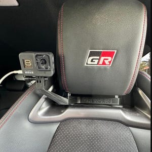 Könnte beinhalten: Eine schwarze GoPro-Kamera, die an einer Kopfstützenhalterung in einem Auto befestigt ist. Die Kopfstütze hat eine schwarze Lederhülle mit roten Nähten und ein GR YARIS-Logo.