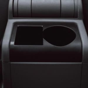 May include: A black car console with a square storage compartment and a circular cup holder. The console is made of a matte black plastic and is positioned between two seats. The top of the console has two rectangular slots.