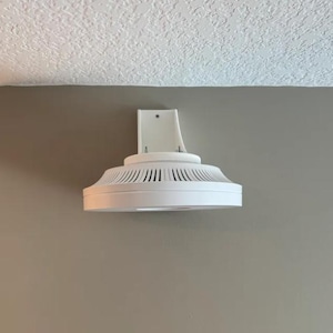 Può includere: Un dispositivo bianco montato a soffitto con un design circolare e una serie di piccoli fori attorno al bordo. Il dispositivo è montato su una parete bianca.