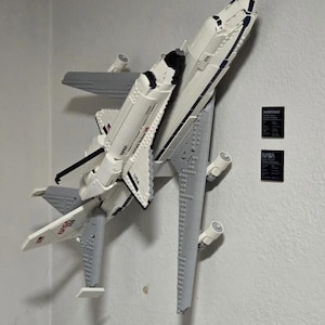 May include: A white and black LEGO space shuttle model mounted on a gray LEGO airplane. The model has the word "NASA" and "USA" printed on it. Two small black plaques with text are visible.