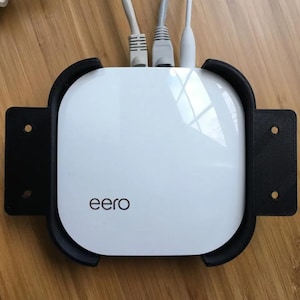 Puede incluir: Un router eero blanco con un soporte de pared negro. El router tiene un logotipo que dice "eero" y está conectado a tres cables Ethernet.