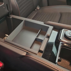 May include: A black plastic organiser with multiple compartments is installed in the centre console of a car. The organiser is designed to hold small items like phones, keys, and other accessories.