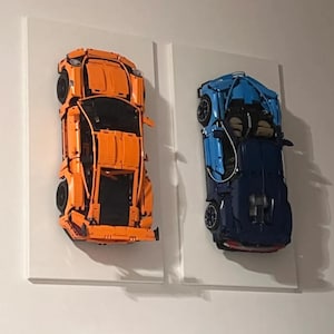 May include: Two Lego car models mounted on white canvases. The car on the left is orange and the car on the right is blue.