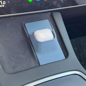 May include: A gray plastic holder for a small white device, likely a wireless earbud case, is mounted in the center console of a car. The holder is designed to fit snugly and securely.