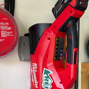 Peut inclure: Une lampe de travail Milwaukee FUEL rouge et noire avec une batterie attachée. La lampe est fixée au mur, à côté d'une bobine de fil de coupe-bordure. Le logo REI Co-op est visible sur la lampe.