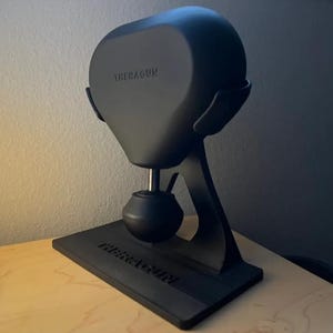 May include: A black Theragun massage gun stand with a round base and a curved arm. The stand has the word "Theragun" printed on the base.