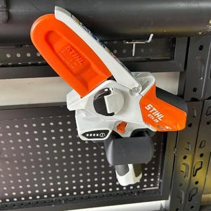 May include: A cordless STIHL GTA 26 battery-powered garden pruner. The tool is white and orange with the STIHL logo. The pruner is hanging on a black metal pegboard.