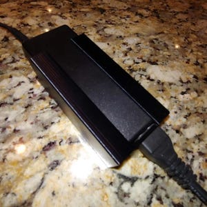 May include: A black rectangular electronic device with a power cord attached. The device has a matte finish and a ridged design on the top. The power cord is black and plugged into the device.