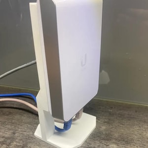 Op de afbeelding: Een wit draadloos toegangspunt met een zilveren rand, gemonteerd op een witte standaard. Het toegangspunt heeft een logo met de tekst "Ubiquiti" aan de voorkant. Drie ethernetkabels zijn aangesloten op het toegangspunt.