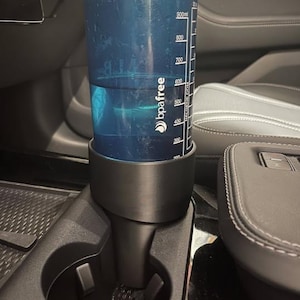 May include: A blue BPA-free water bottle, partially filled with water, sits in a black car cup holder. The bottle has measurement markings up to 900ml. The cup holder is integrated into the car's console, with black and gray leather seats visible in the background.