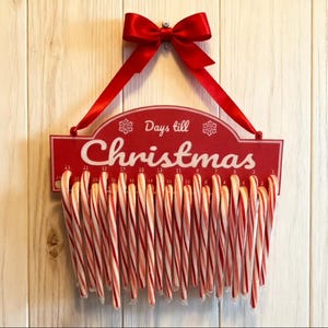 Könnte beinhalten: Ein rotes Holz-Weihnachts-Countdown-Schild mit den Worten "Days till Christmas" und einer Reihe von Zuckerstangen, die darunter hängen. Oben befinden sich ein rotes Band und eine Schleife. Das Schild hängt vor einem hellen Holzhintergrund.