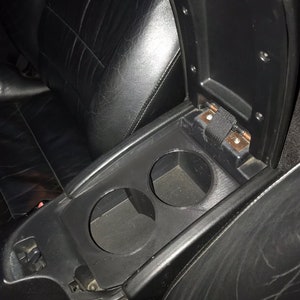 Puede incluir: Un interior de coche negro con una consola central que tiene dos portavasos y un compartimento de almacenamiento. El compartimento de almacenamiento está abierto y tiene una correa negra.