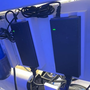 May include: Two black power adapters with green indicator lights are mounted on a white surface. Black cords are neatly coiled. The setup suggests an electronic device or system, possibly for aquariums or similar applications.