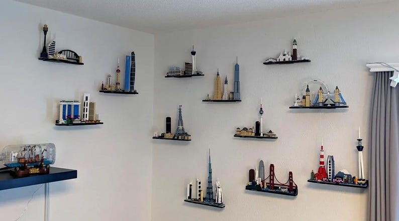 Puede incluir: Una pared blanca con 11 estantes negros que muestran paisajes urbanos en miniatura de Lego. Los modelos de Lego incluyen la Torre Eiffel, el Empire State Building, el London Eye y la &Oacute;pera de Sydney.