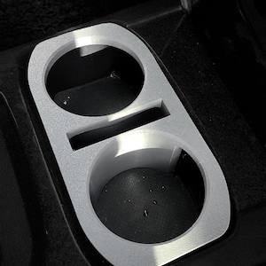 May include: Silver-colored car cup holder with two circular openings and a rectangular slot. The cup holder is designed to fit into a car's center console. The surface has a textured appearance.