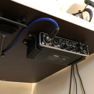 Könnte beinhalten: Ein schwarzes Audio-Interface mit Knöpfen und Reglern ist unter einem Schreibtisch montiert, an dem ein blaues Kabel angeschlossen ist.