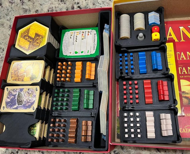 May include: A board game organizer with various game pieces, including cards, dice, and wooden tokens in multiple colors. The game pieces are neatly arranged in black trays within a red box, ready for play.
