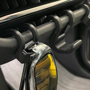 May include: Black car vent hooks designed to hold keys or other small items. The hooks are attached to a car vent and are made of a durable material. A car key is hanging from one of the hooks.