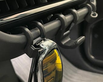 Op de afbeelding: Zwarte autoventielhaken ontworpen om sleutels of andere kleine items op te hangen. De haken worden aan een autoventiel bevestigd en zijn gemaakt van een duurzaam materiaal. Een autosleutel hangt aan een van de haken.