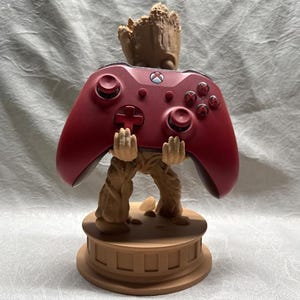 May include: A brown Groot figure holding a red Xbox controller. The figure is on a brown base with a circular design. The controller has the Xbox logo and button details.