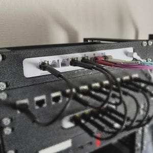 Pode incluir: Close-up de um rack de rede preto com vários cabos conectados a um dispositivo branco. Os cabos são pretos, vermelhos, roxos e azuis. O rack é montado em uma parede.