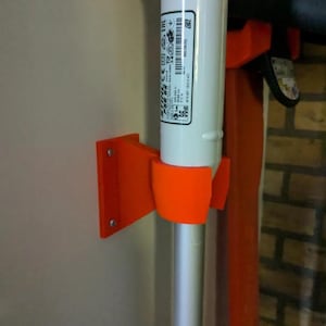 Puede incluir: Un soporte de pared naranja impreso en 3D que sostiene una herramienta de jardinería blanca y plateada. El soporte está fijado a una pared de color claro con tornillos. La herramienta de jardinería tiene una etiqueta con texto y símbolos.