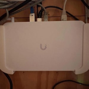May include: A white rectangular device with a U logo mounted on a wooden wall. The device has a white plastic casing and is connected to several cables.